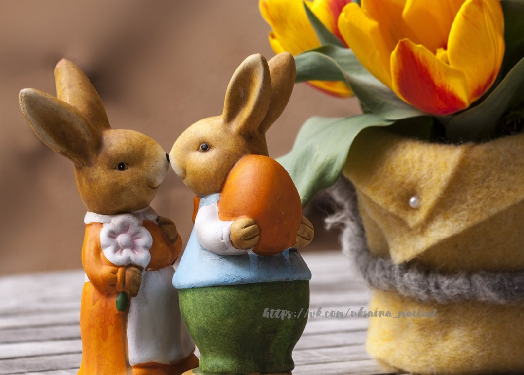 Two ceramic bunny figurines with an orange egg beside tulips in a felt-wrapped pot