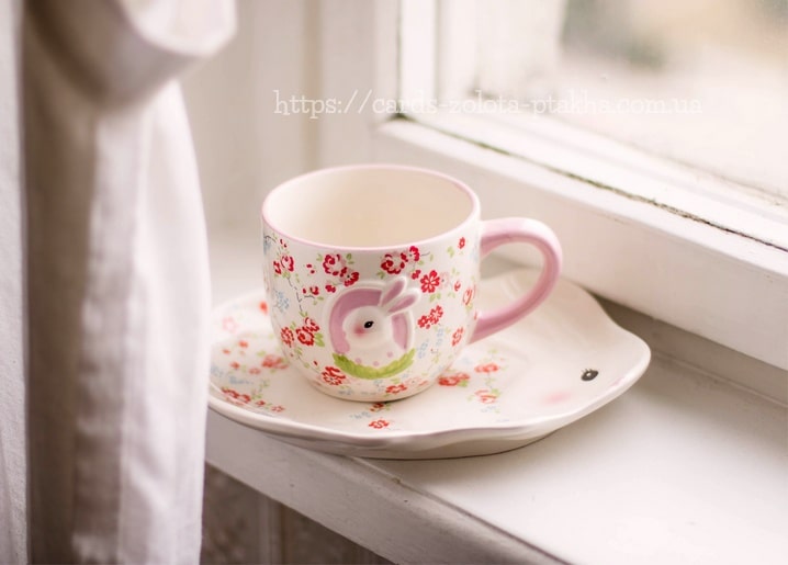 Weiße Tasse mit Hasenmotiv und rosa Blüten auf der Untertasse am Fenster