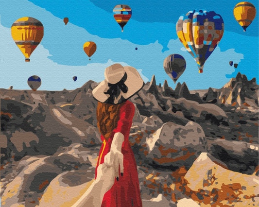 Woman in a red dress and straw hat watching hot air balloons rise over Cappadocia's rock valleys