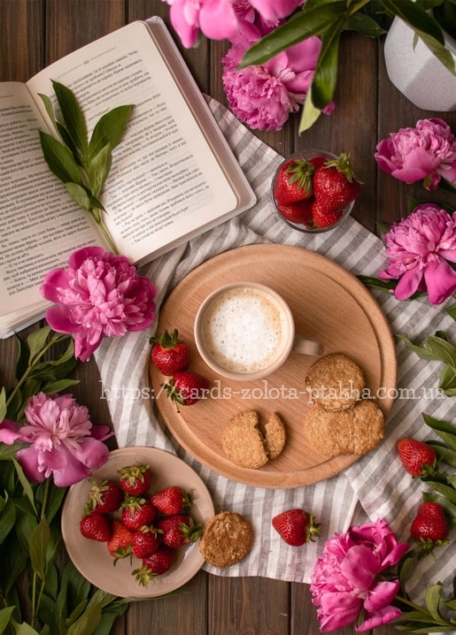Cappuccino, Haferkekse und Erdbeeren auf einem Holzbrett neben einem aufgeschlagenen Buch und rosa Pfingstrosen.