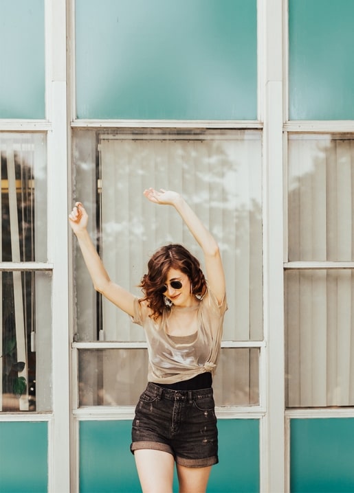 Young woman in sunglasses raises her arms and dances in front of a teal-framed paned window.