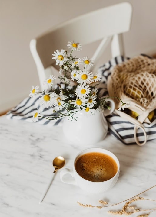 Weißer Krug mit Kamillenblüten neben einer Kaffeetasse auf Marmor