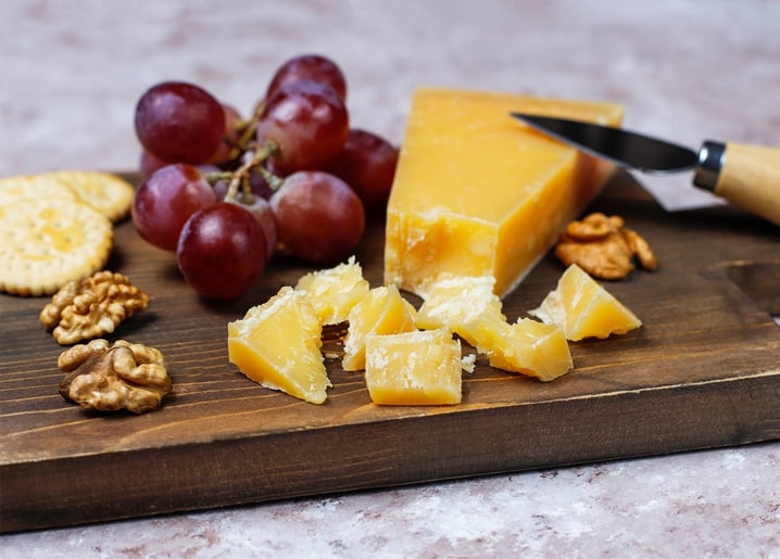 Wedge of cheddar cheese cut into chunks with grapes, walnuts and crackers on a dark wooden board