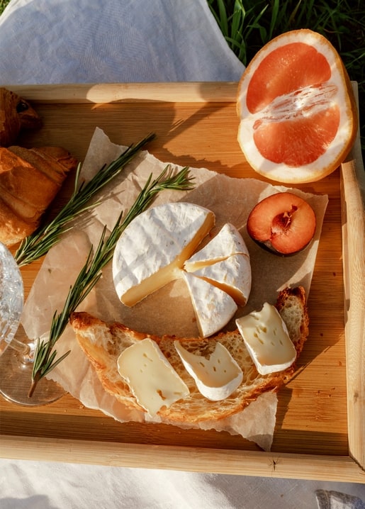 Wooden tray with soft cheese, croissant, rosemary and halved grapefruit in sunlight