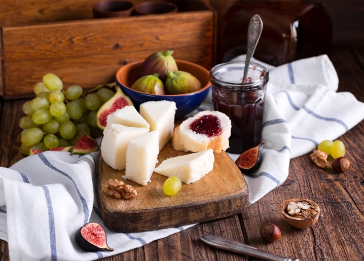 Cheese cubes, fresh figs, grapes and a jar of jam with bread, walnuts and hazelnuts on striped linen