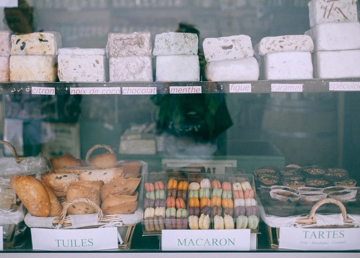 Schaufenster mit beschrifteten Käselaiben und Tabletts voller Macarons, Tuiles und Tartes