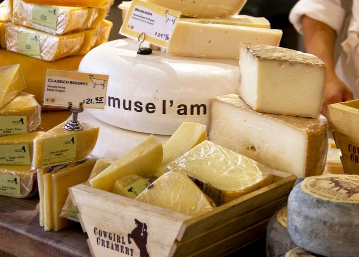 Cheese shop counter with wrapped wedges, handwritten price tags, and a whole wheel behind glass