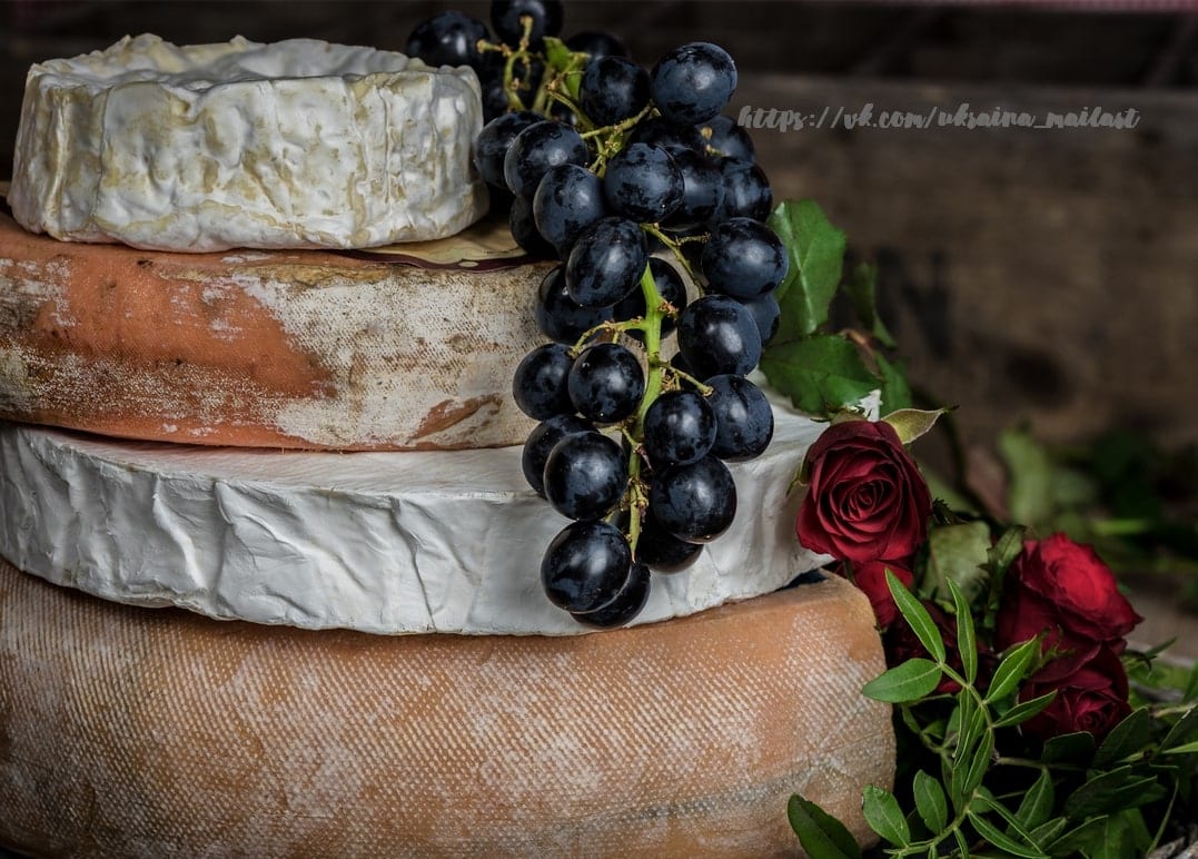Stacked cheese wheels draped with dark grapes and red roses beside them