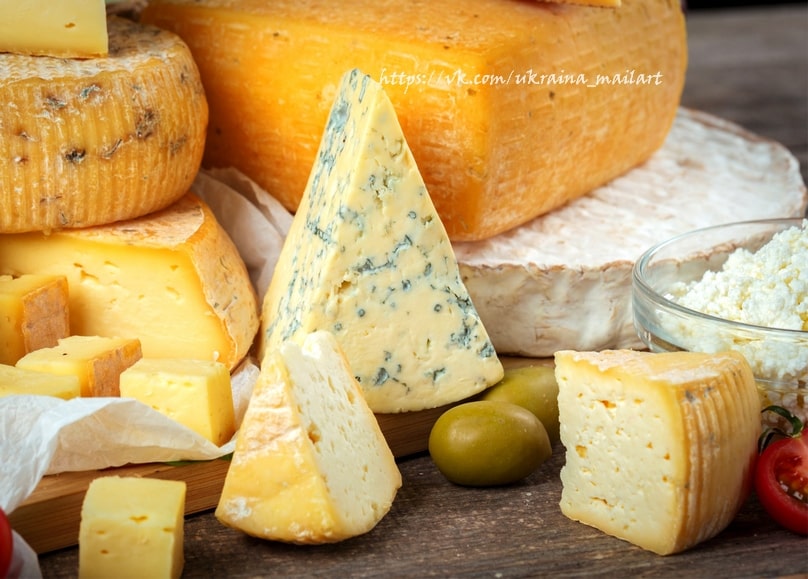 Wheels of cheese, a blue cheese wedge, cottage cheese, and green olives on a wooden table