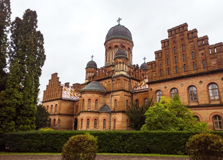 Nahaufnahme der Kuppeltürme und des gemusterten Dachs der Universität Tscherniwzi hinter einer Hecke
