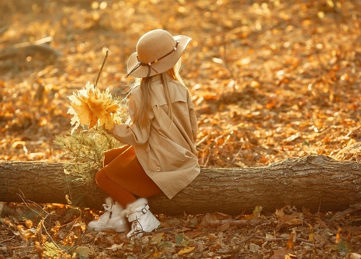 Child in a hat and coat sitting on a log, holding autumn leaves