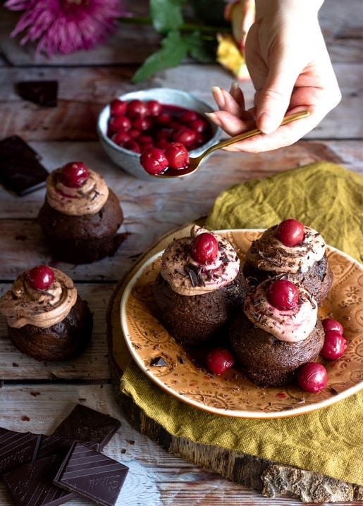 Chocolate cupcakes topped with cream and cherries, a spoon adding syrup, on a rustic wood table.