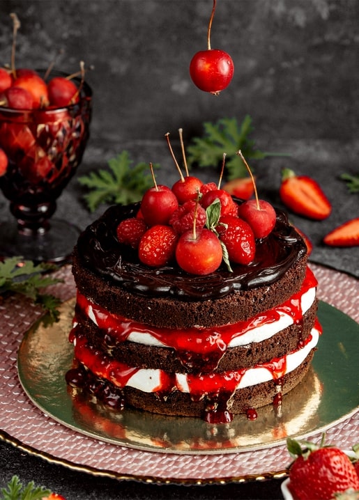 Chocolate drip cake topped with cherries, raspberries and strawberries, one cherry falling
