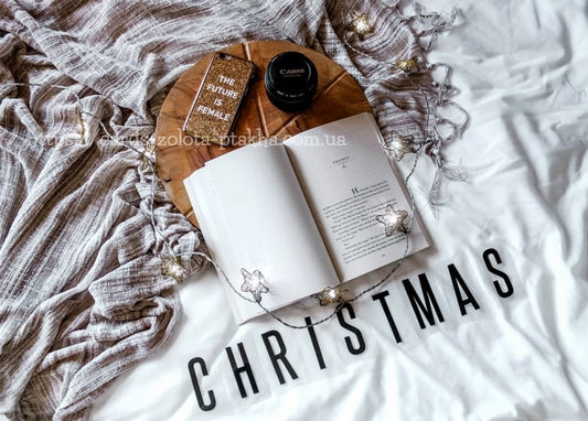 Aufgeschlagenes Buch, Handyhülle und Objektivdeckel neben Sternenlichterkette und Schriftzug CHRISTMAS