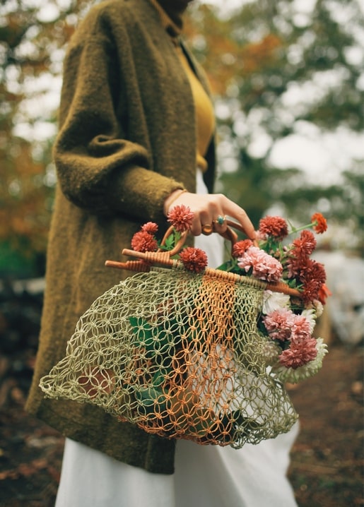 Woman in a wool cardigan holding a mesh bag full of pink and coral chrysanthemums.