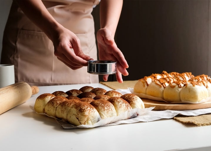 Hände bestäuben frisch gebackene Brötchen mit Zimt neben einem Nudelholz auf der Küchentheke.