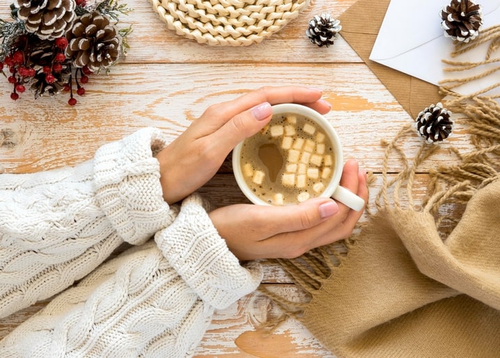Hände im Strickpullover halten eine Tasse Kakao mit Marshmallows neben Zapfen und Kraftpapier-Umschlag