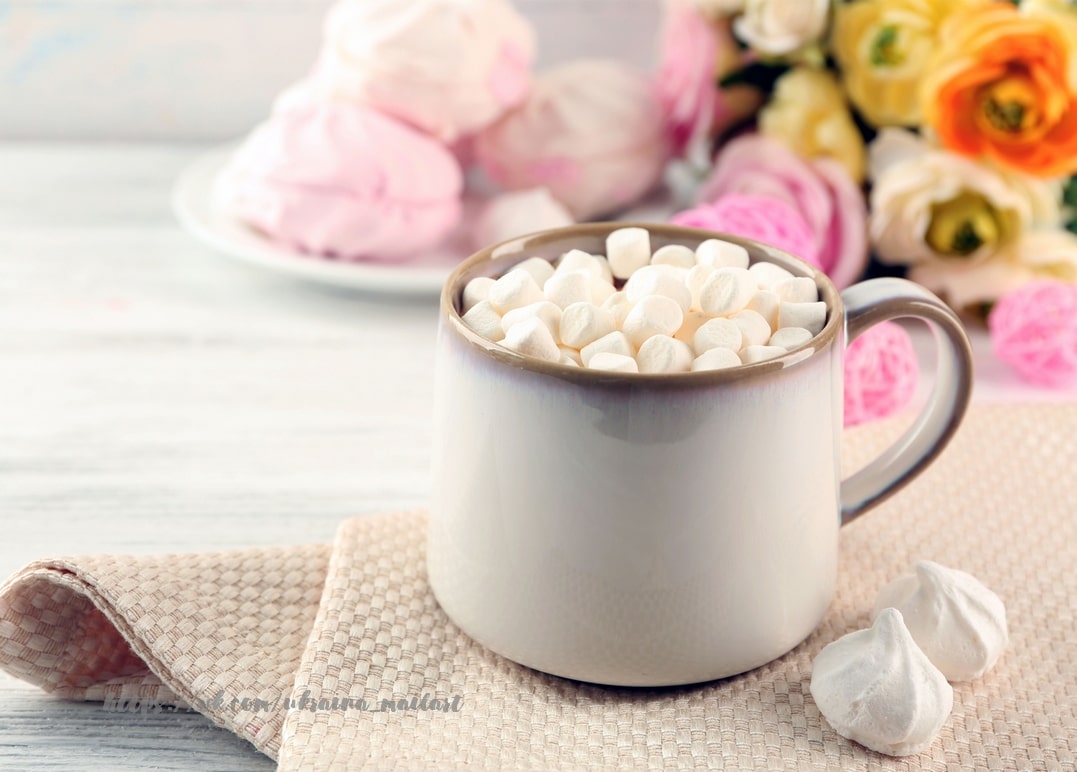Tasse Kakao mit Mini-Marshmallows neben rosa-weißem Baiser