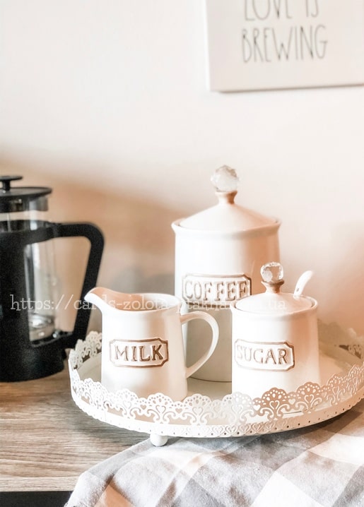 Weiße Keramikdosen für Kaffee, Zucker und Milch auf einem Spitzentablett neben einer French Press.