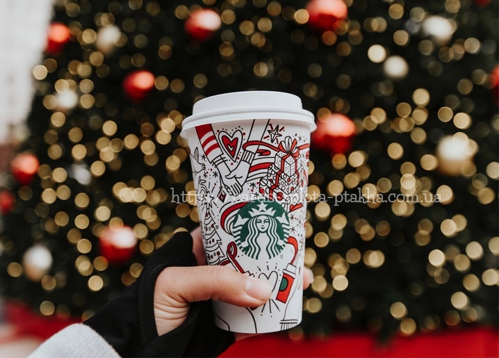 Hand hält einen festlichen Kaffeebecher vor einem Weihnachtsbaum mit roten Kugeln und Bokeh-Licht