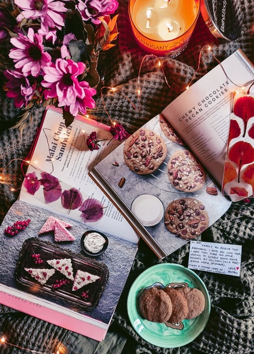 Open cookbook with chocolate pecan cookies, pink flowers, and candlelight on a cozy blanket