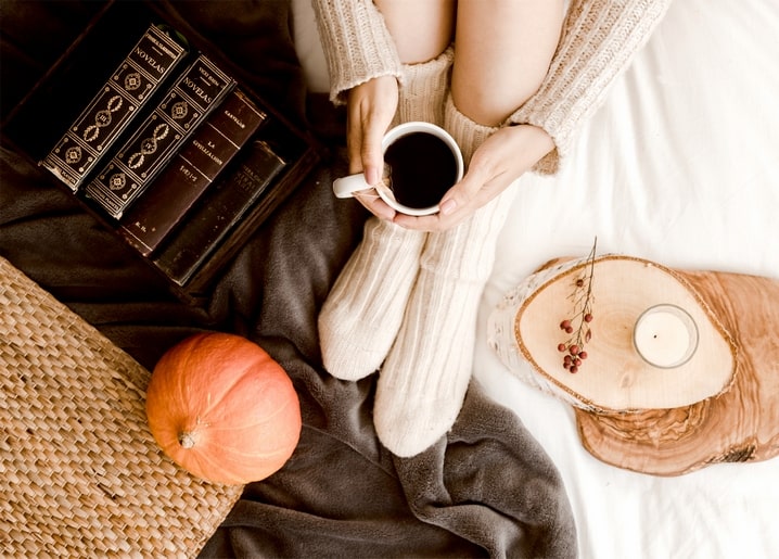 Woman in wool socks holding a coffee mug beside a pumpkin, books and a candle on linen
