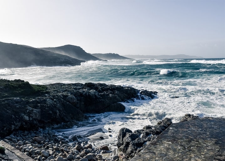Meereswellen brechen über dunklen Felsen einer nebligen, bergigen Küste