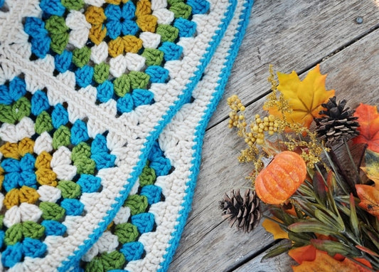 Teal and mustard granny-square crochet blanket beside a pumpkin, pinecones, and maple leaves on wood