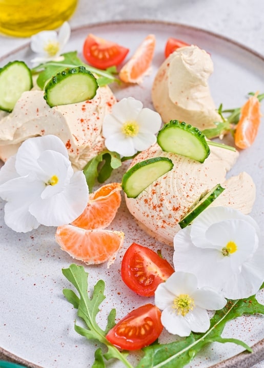 Teller mit Frischkäse, Gurken, Kirschtomaten, Mandarine und essbaren Blüten