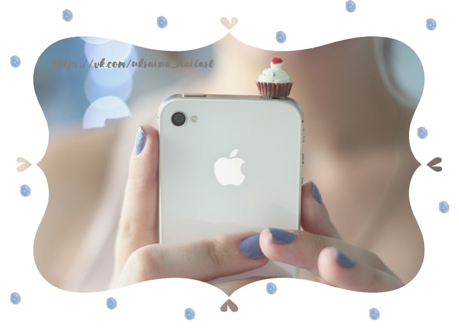 White smartphone with a tiny cupcake charm on top, held by hand with blue nail polish