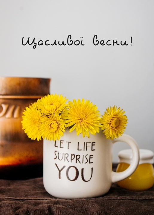 Букет жовтих кульбаб у чашці з написом 'Let Life Surprise You', поруч баночка меду та мідна бляшанка.
