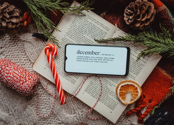 Open book with a phone showing the word december, candy canes, pinecones and a dried orange slice on a knit blanket