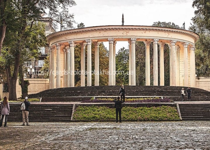 Circular colonnade on a stone terrace above the Dnipro riverside promenade, with visitors and autumn trees.