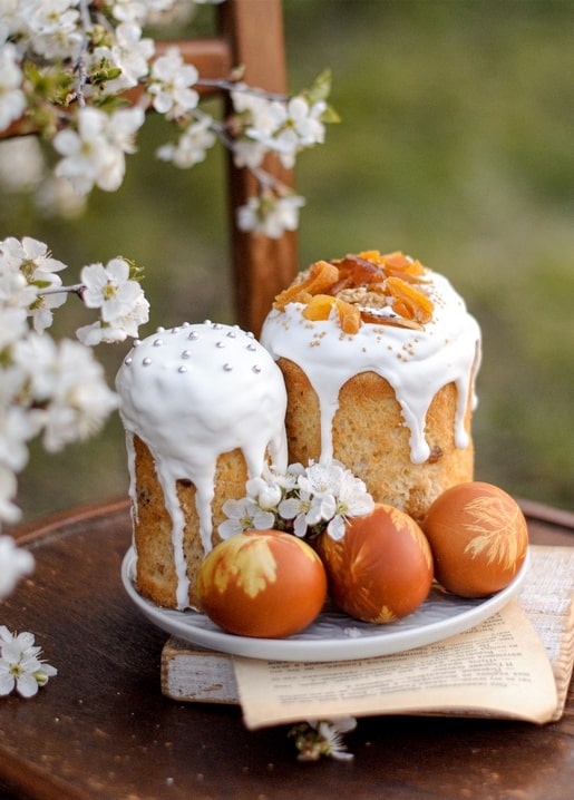 Two paska cakes with white icing and painted orange eggs among cherry blossom on a wooden chair