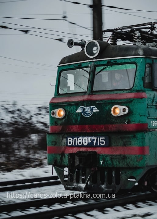 Green electric locomotive with headlights on, moving through snow on Ukrainian railway tracks