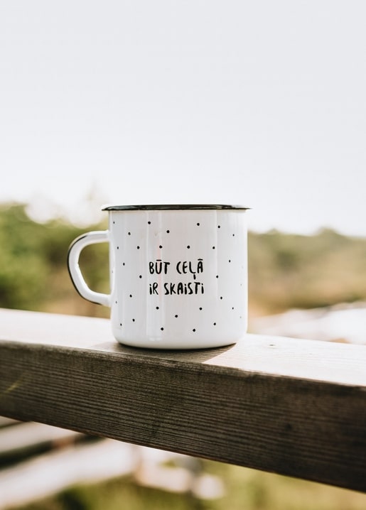 Weißer Emaille-Becher mit handgeschriebenem Spruch auf einem verwitterten Holzgeländer