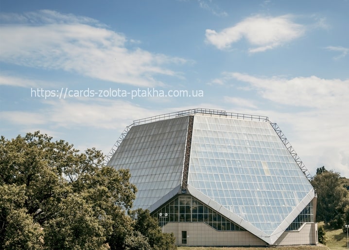 Faceted glass building with a diamond-shaped roof on a green hill among oak trees under a blue sky