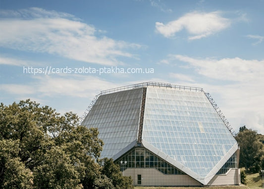 Faceted glass building with a diamond-shaped roof on a green hill among oak trees under a blue sky