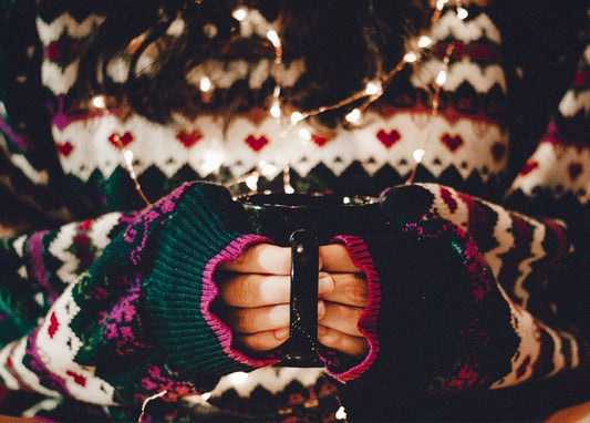 Hands in a fair isle sweater holding a mug, wrapped in glowing fairy lights