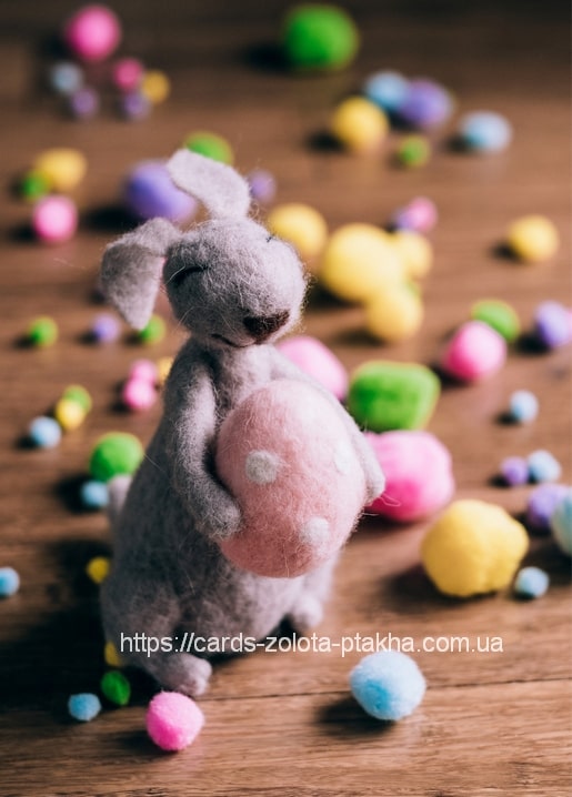 Needle-felted gray bunny holding a pink polka-dot egg among colorful wool pom-poms