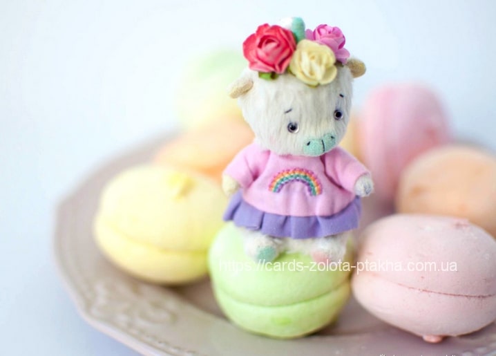 Felted lamb in a rainbow sweater and flower crown sitting on pastel macarons