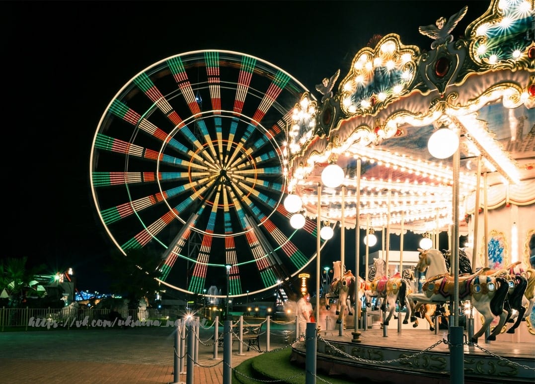 Beleuchtetes Riesenrad dreht sich neben einem hell erleuchteten Karussell mit Pferden bei Nacht