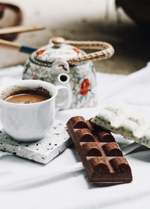 Blumen-Porzellan-Teekanne neben einer Kaffeetasse und Riegeln dunkler und weißer Schokolade