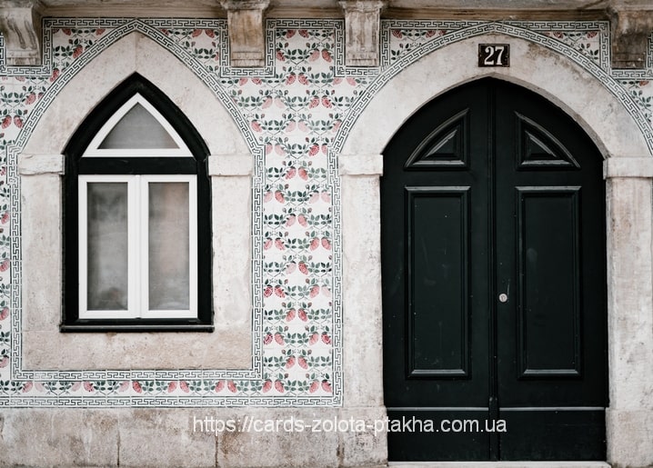 Weiße Fassade mit rosa Blumenkacheln, spitzbogigem Fenster und schwarzer Flügeltür Nr. 27.