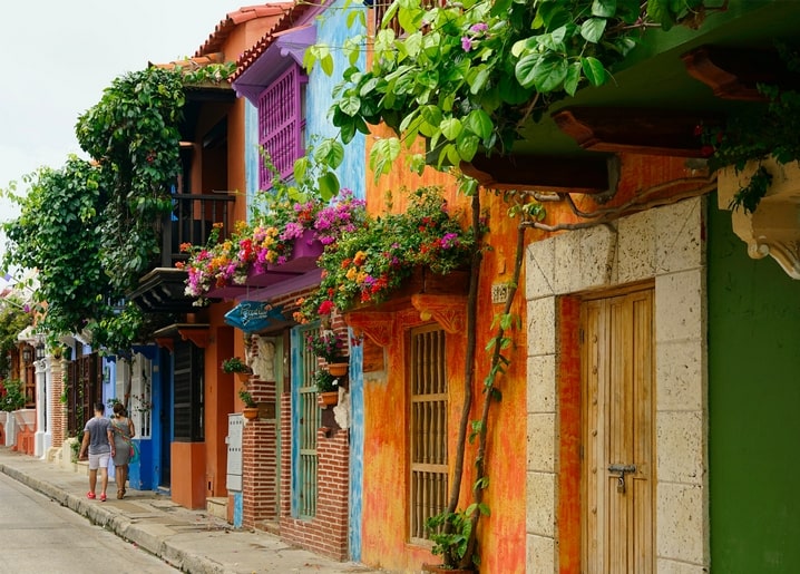 Bunte Kolonialgasse mit blühenden Balkonen, orangefarbenen und türkisen Häusern und einem spazierenden Paar.