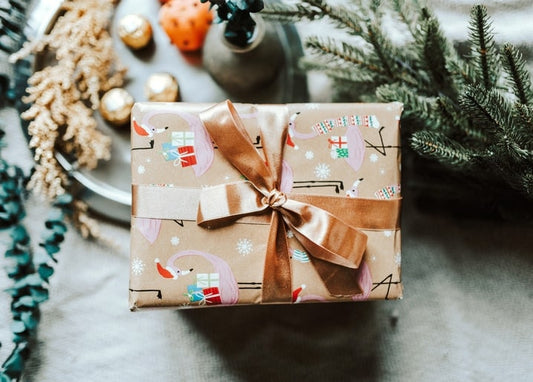 Gift wrapped in patterned kraft paper with a gold ribbon bow among pine branches