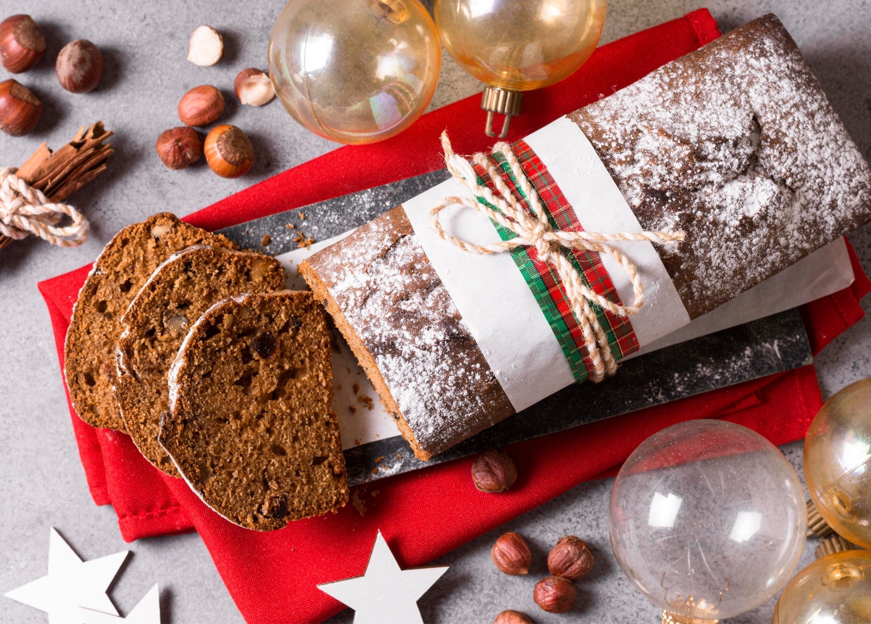 Aufgeschnittenes Lebkuchenbrot mit Puderzucker und Karoband zwischen Haselnüssen und Glaskugeln