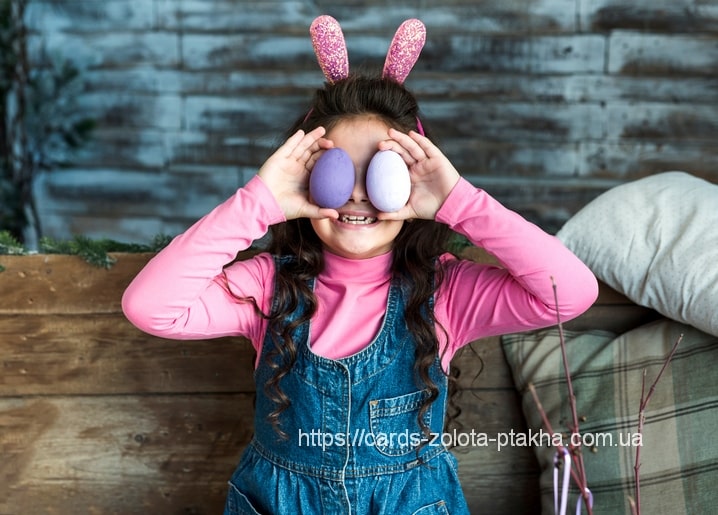 Girl in pink bunny ears holding two lilac Easter eggs over her eyes