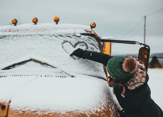 Girl in a pompom hat drawing a heart in snow on a truck windshield