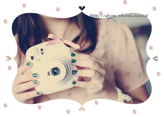 Mädchen mit grün lackierten Nägeln hält eine weiße Fujifilm-Instax-Kamera mit rosa Schleife
